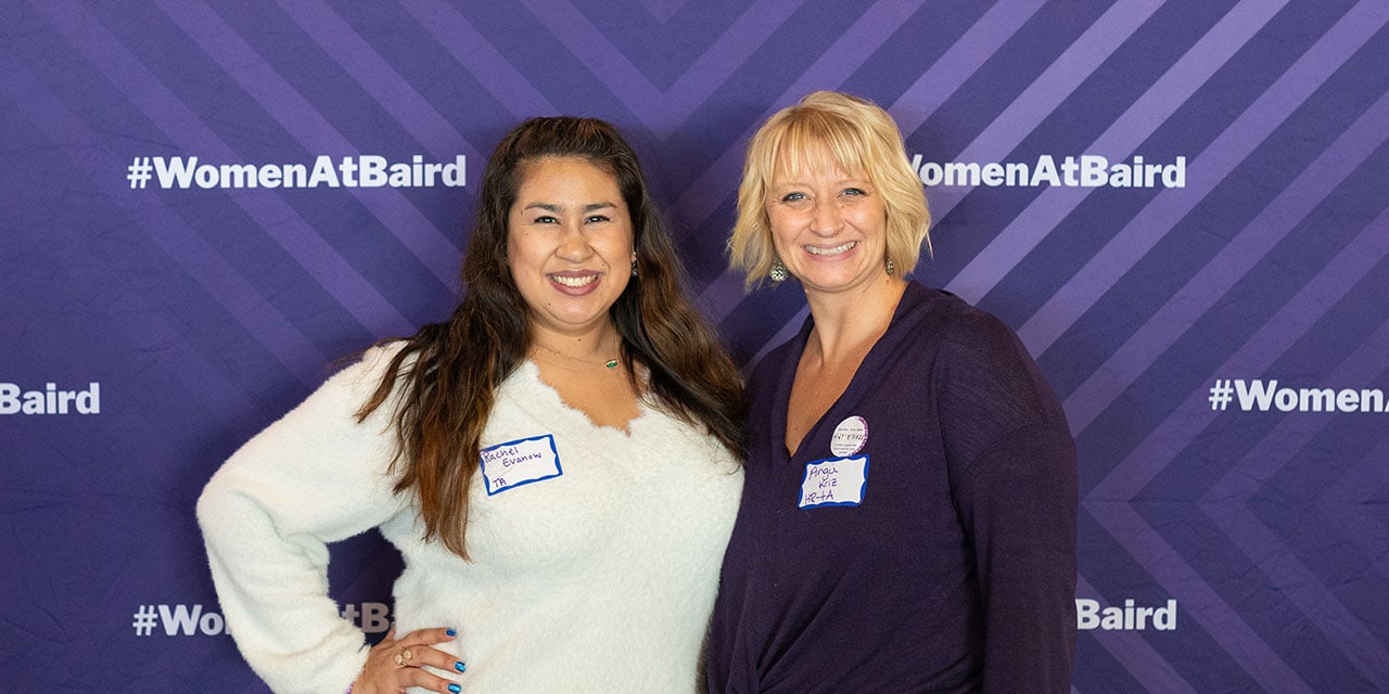 Two SilverPeak Strategic Capital associates in front of a purple banner with #womenatbaird hashtags